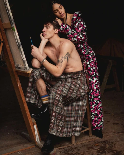 A man in a kilt sits by an easel with a painting, while a woman in a floral dress stands behind him in a dimly lit indoor setting.