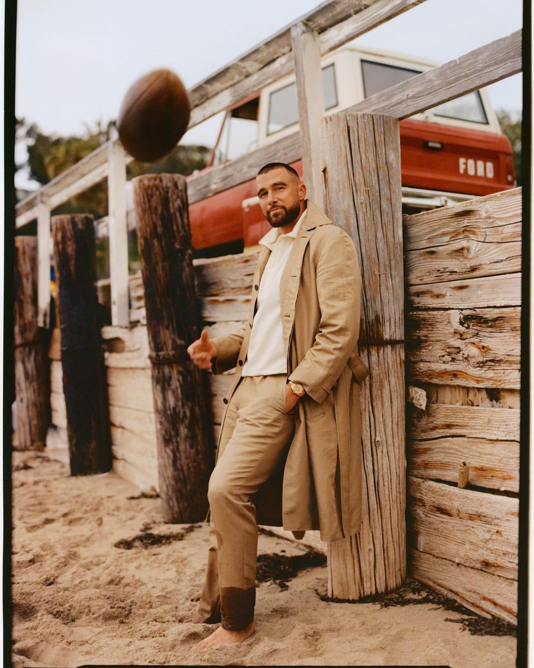 Adult male wearing tan-colored trench coat, pants, and white top leaning against wood poles with a football in the air and a red truck behind.