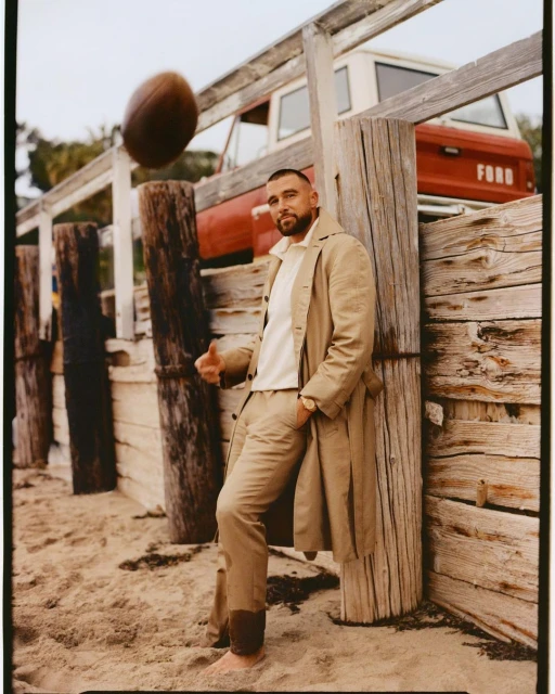 Adult male wearing tan-colored trench coat, pants, and white top leaning against wood poles with a football in the air and a red truck behind.