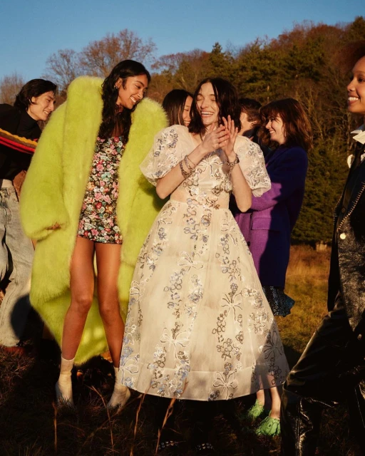 Group of stylish women in floral dresses and fashionable outerwear, posing together in a sunny, grassy outdoor setting for a fashion editorial.