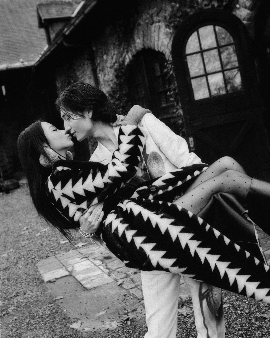 A couple embraces in front of a stone building, the woman in a patterned coat and the man in a light jacket in a black and white photograph.