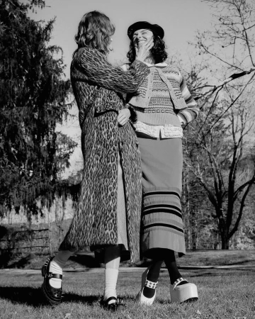 Two women pose outdoors in leopard-print coat, knit sweater, and platform shoes. Trees and a grassy lawn serve as the backdrop.