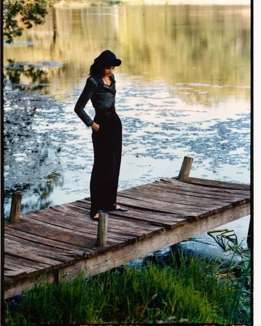 An adult woman stands on a rustic wooden pier with a lake and trees in the background, wearing wide-legged pants and a hat, posing thoughtfully.