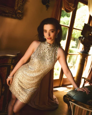 Woman in a gold sequin sleeveless dress posing indoors by a large window with draped curtains and antique furniture.