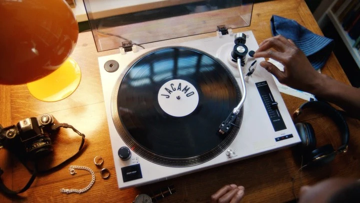 Overhead shot of a vinyl record player on a wooden table, with a person's hand visible, along with accessories and a lamp providing warm lighting.