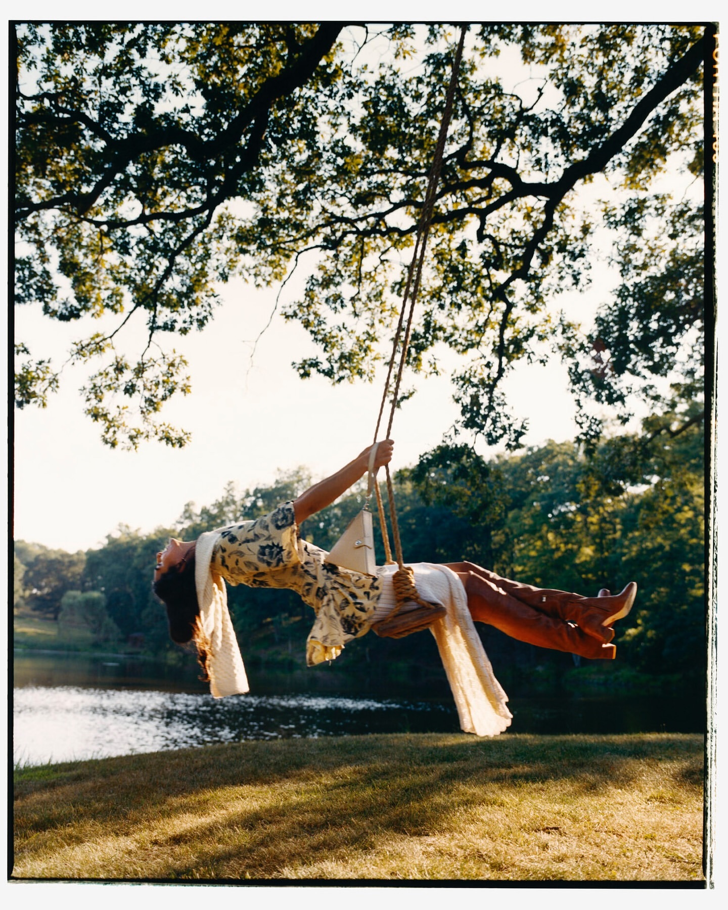 Woman in dress swings on tree swing over water. Outdoor nature scene with large tree, green grass, lake and wooded background.