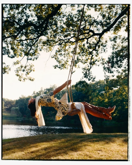 Woman in dress swings on tree swing over water. Outdoor nature scene with large tree, green grass, lake and wooded background.