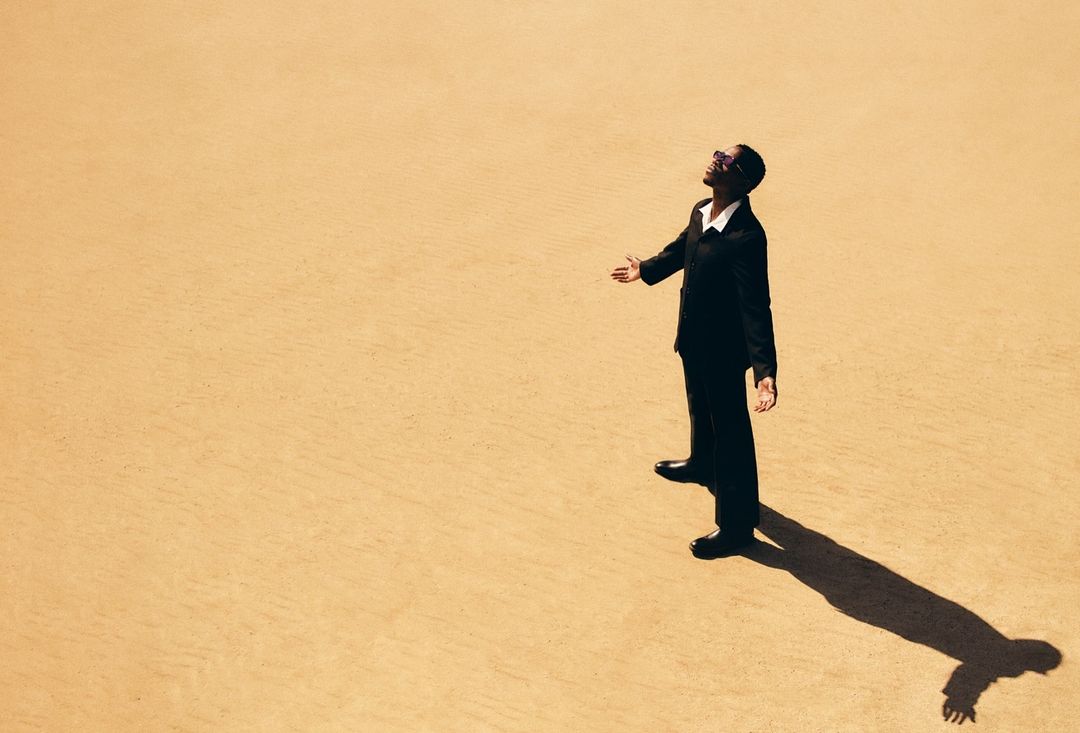 Adult man in business suit stands in desert looking up with arms outstretched; an overhead shot highlights shadow on sand.
