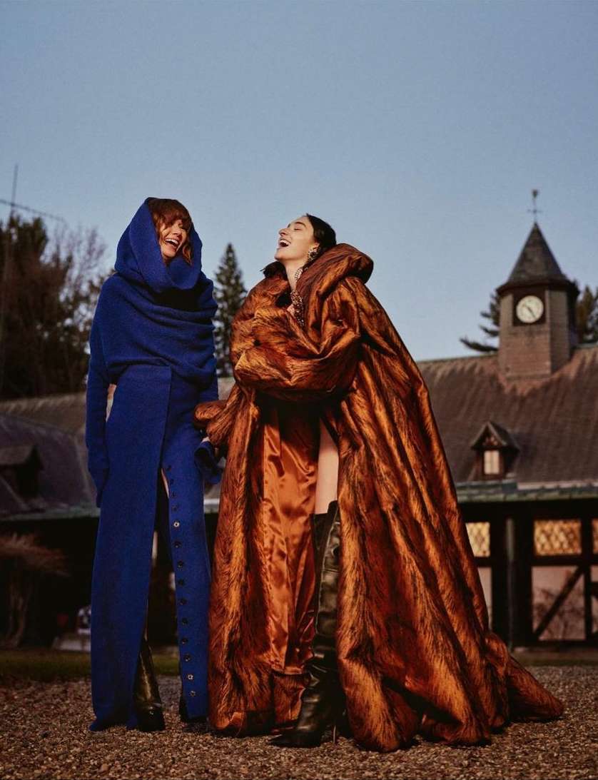 Two adult females in striking blue and copper-colored hooded coats are outdoors, laughing, with a building in the background on an overcast day.