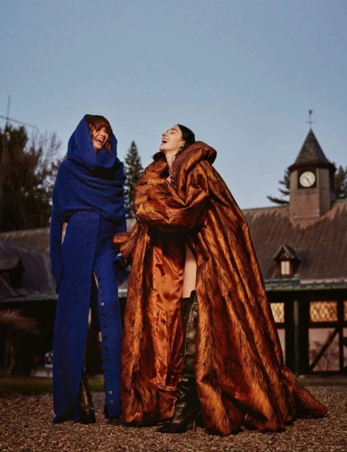 Two adult females in striking blue and copper-colored hooded coats are outdoors, laughing, with a building in the background on an overcast day.