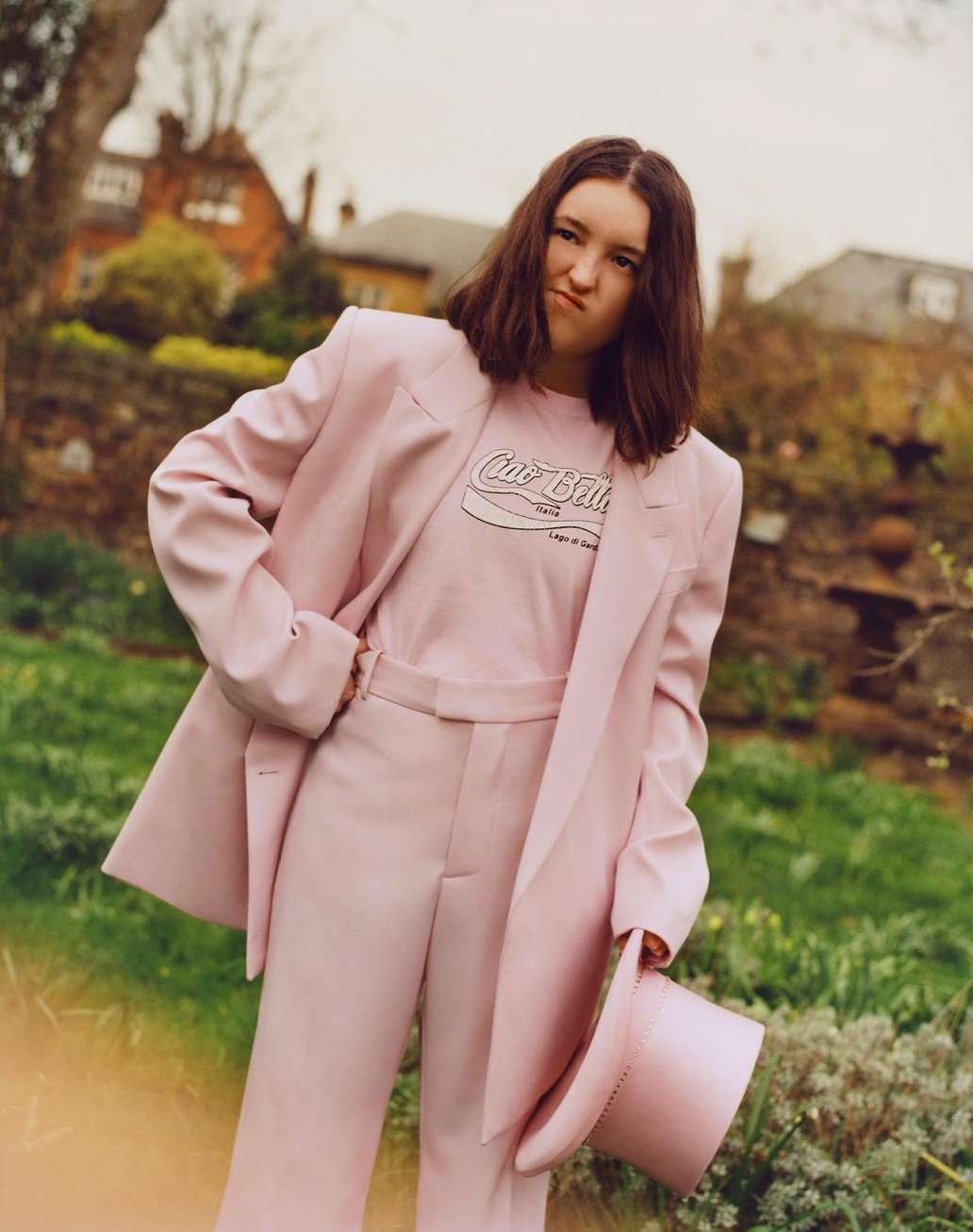 Young woman in a pink suit strikes a playful pose outdoors in a garden setting with plants, flowers, and a house in the background.