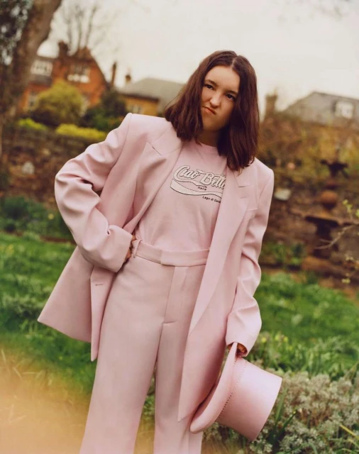 Young woman in a pink suit strikes a playful pose outdoors in a garden setting with plants, flowers, and a house in the background.