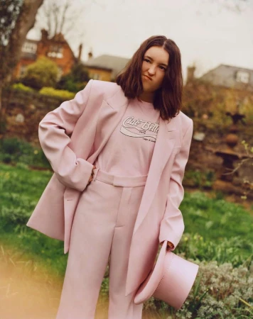 Young woman in a pink suit strikes a playful pose outdoors in a garden setting with plants, flowers, and a house in the background.