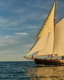 A person sitting on the bowsprit of a large dark wooden sailboat with cream sails navigating choppy open ocean under a partly cloudy blue sky