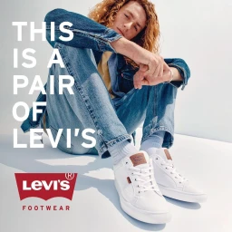 Young man with curly red hair wearing a denim jacket and jeans, seated against a white background with white sneakers.