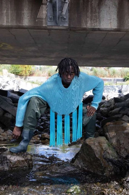 A person with dark skin and dreadlocks poses by a stream under a bridge, wearing a blue knit sweater with decorative fringe and boots.