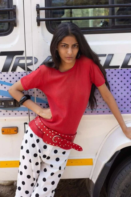 Woman in red top and polka-dot pants leaning against a white vehicle, posing outdoors with a serious expression and long black hair.
