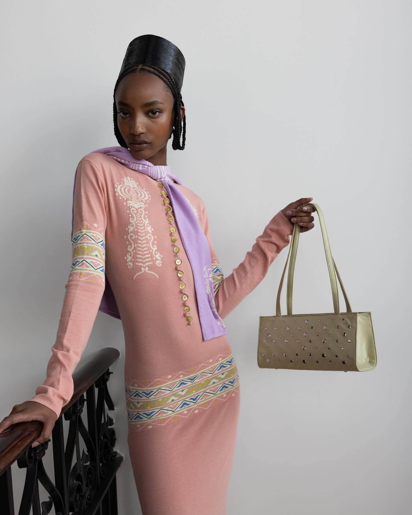 Fashionable woman in pink dress and lilac scarf holds gold purse in studio setting with black railing, showcasing modern style.