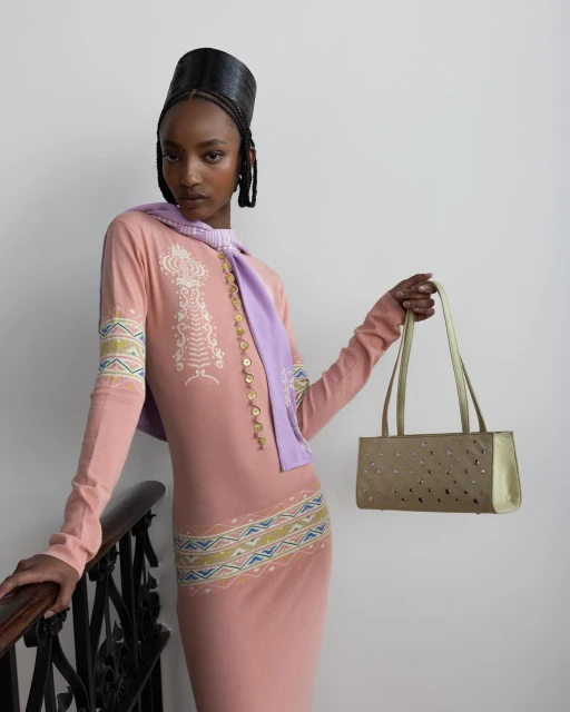 Fashionable woman in pink dress and lilac scarf holds gold purse in studio setting with black railing, showcasing modern style.