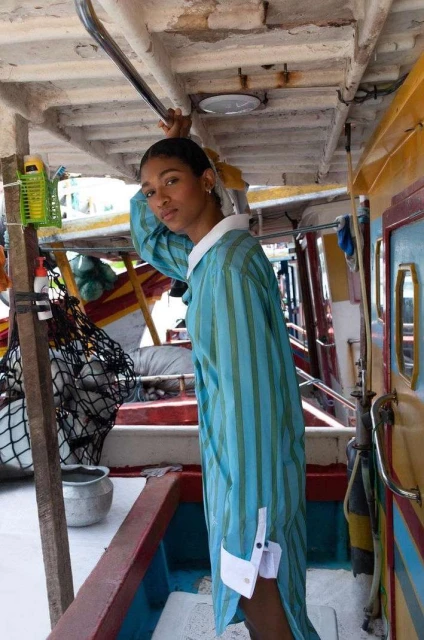Elegant woman in striped blue dress with white collar and cuffs poses on a boat with a weathered interior, looking at the camera.