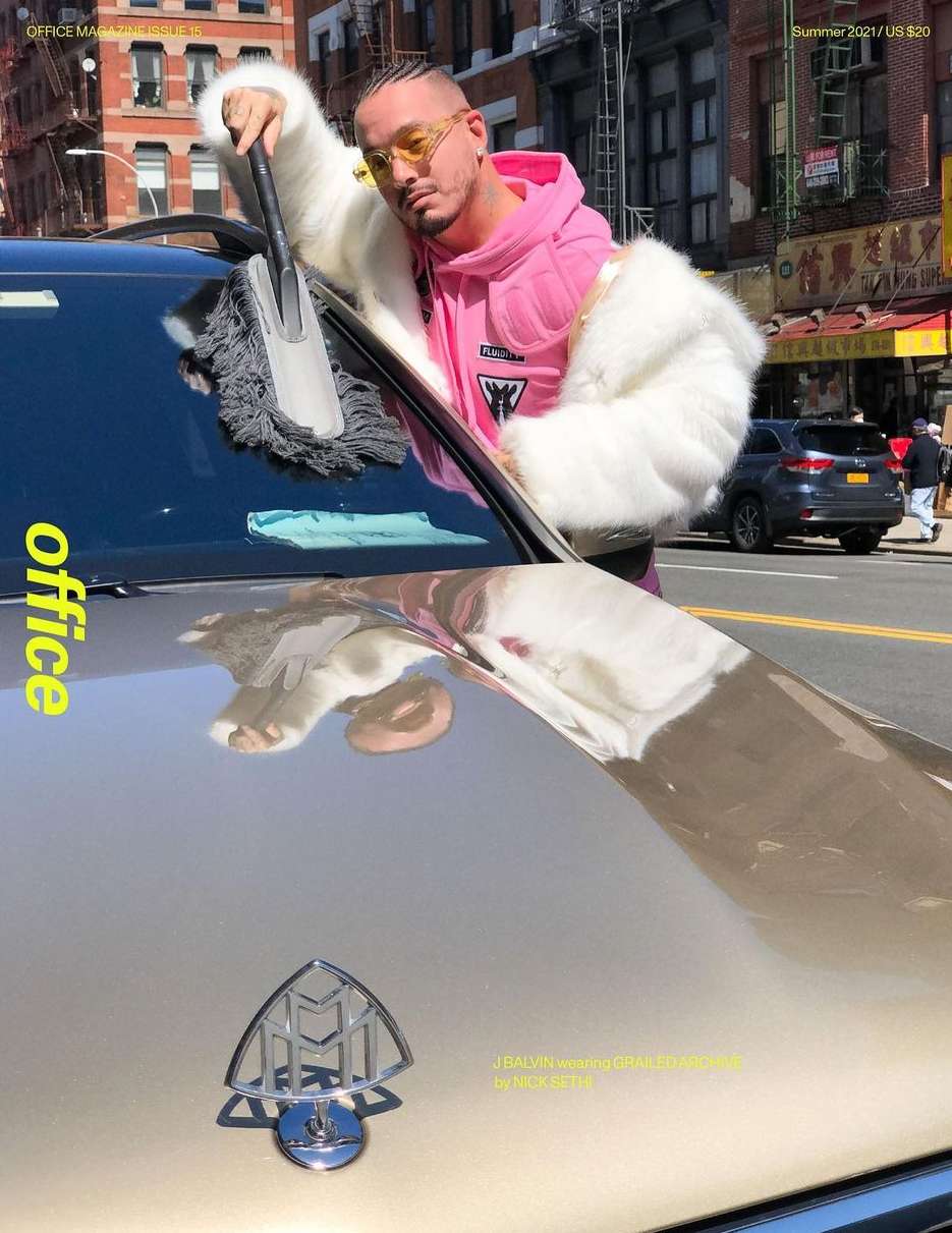 Singer Maluma poses with a duster on the hood of a car in an urban setting for a magazine cover, wearing a fur coat and yellow sunglasses.
