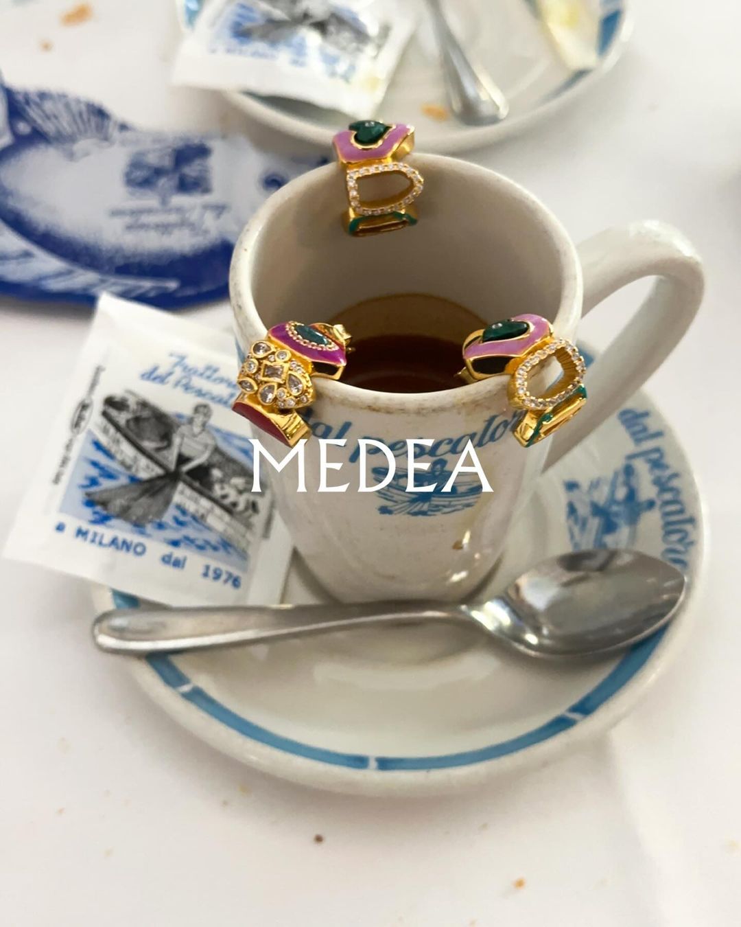 Close-up of coffee cup with ornate letter-shaped rings. The rings are encrusted with jewels and gold. MEDEA logo is visible on the cup.