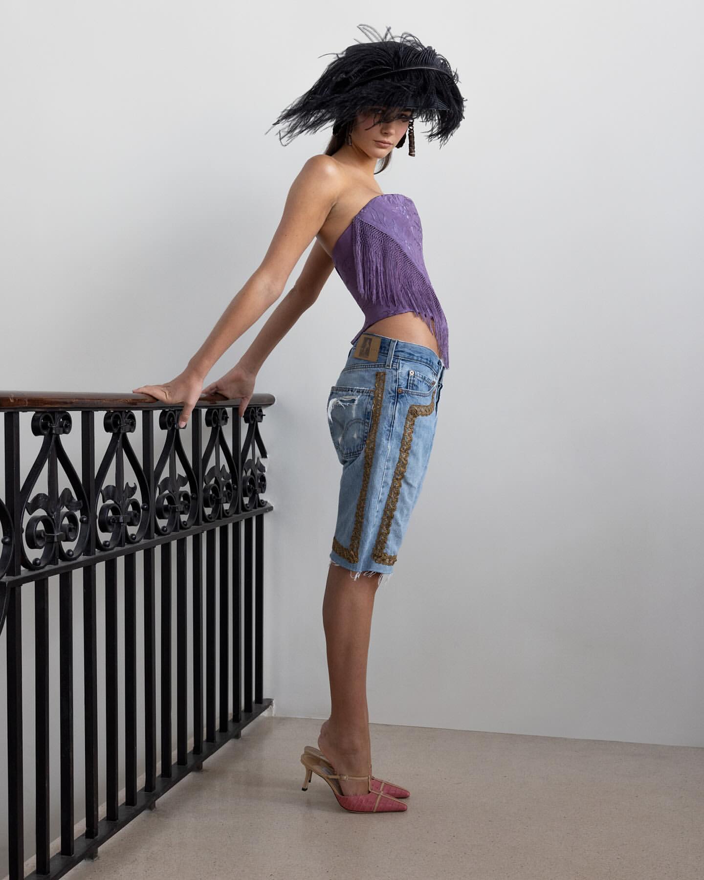 A fashion model poses indoors wearing a purple top, jeans, high heels, and a feathered hat while leaning on a black metal railing.