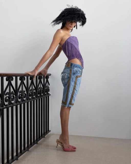 A fashion model poses indoors wearing a purple top, jeans, high heels, and a feathered hat while leaning on a black metal railing.