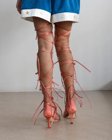 Rear view of legs wearing coral pink lace up gladiator heels and blue shorts with star embroidery against a plain studio background.