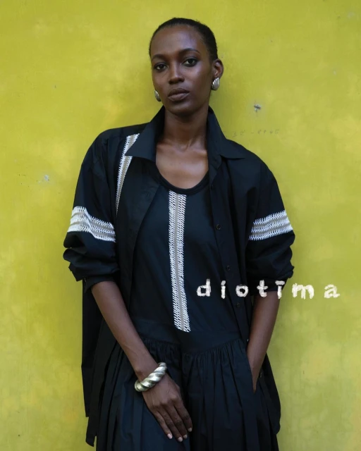 A Black woman poses against a yellow wall, wearing a black dress and open shirt with silver bracelet and earrings in a medium shot.