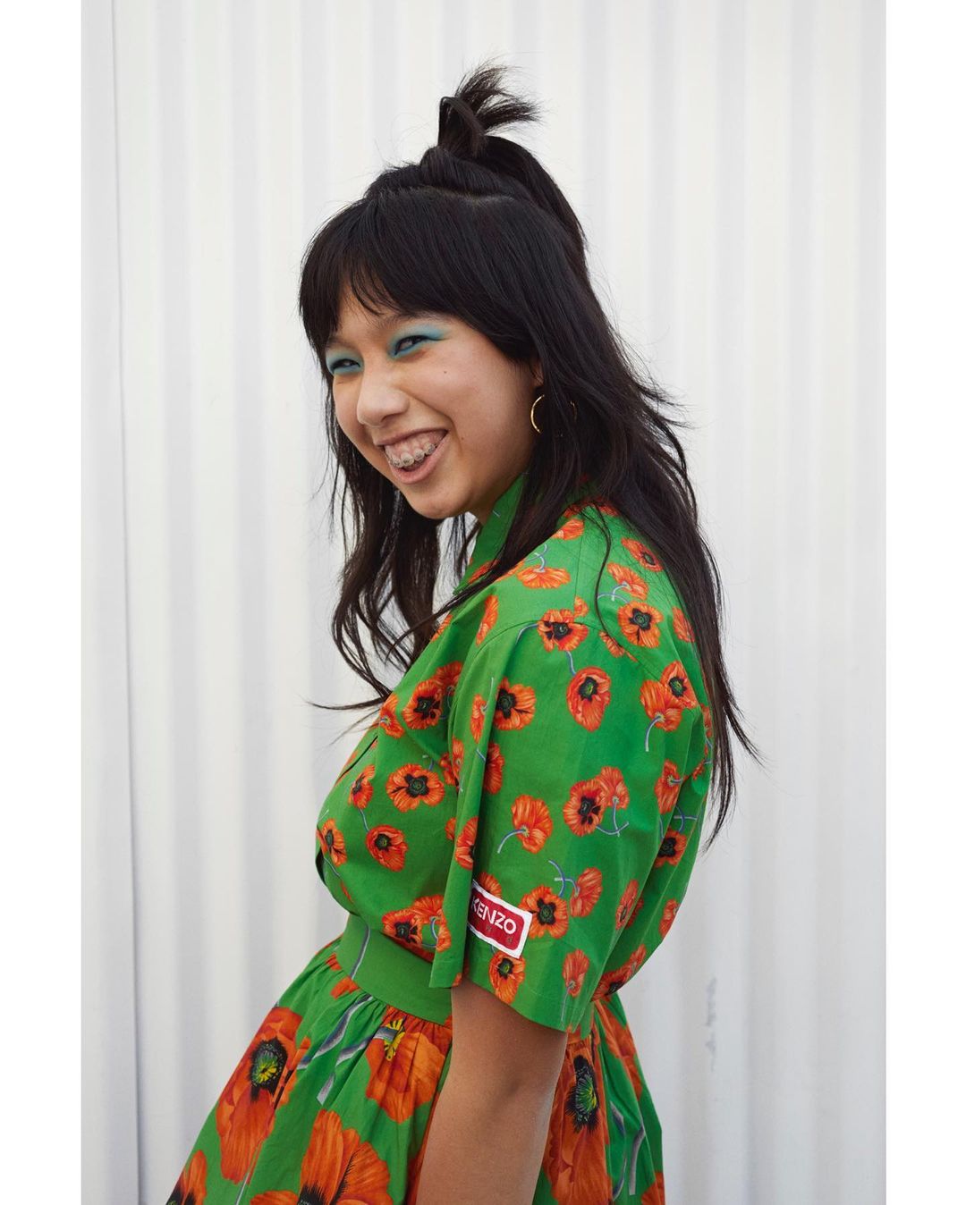 Smiling Asian woman in a green floral dress with red poppies, bright blue eyeshadow, and braces. She is posing in front of a white wall.