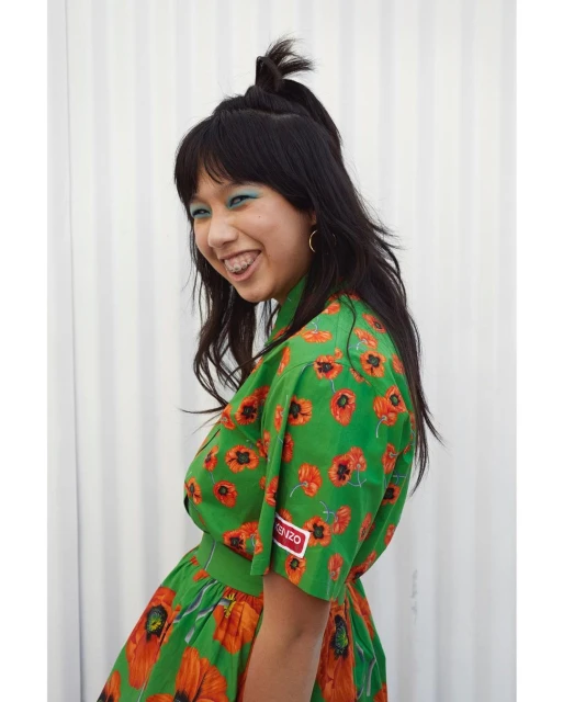 Smiling Asian woman in a green floral dress with red poppies, bright blue eyeshadow, and braces. She is posing in front of a white wall.