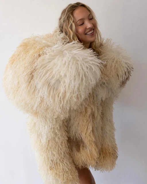 Portrait of a smiling adult woman with wavy blonde hair wearing a large, fluffy, cream-colored fur coat against a plain white background.