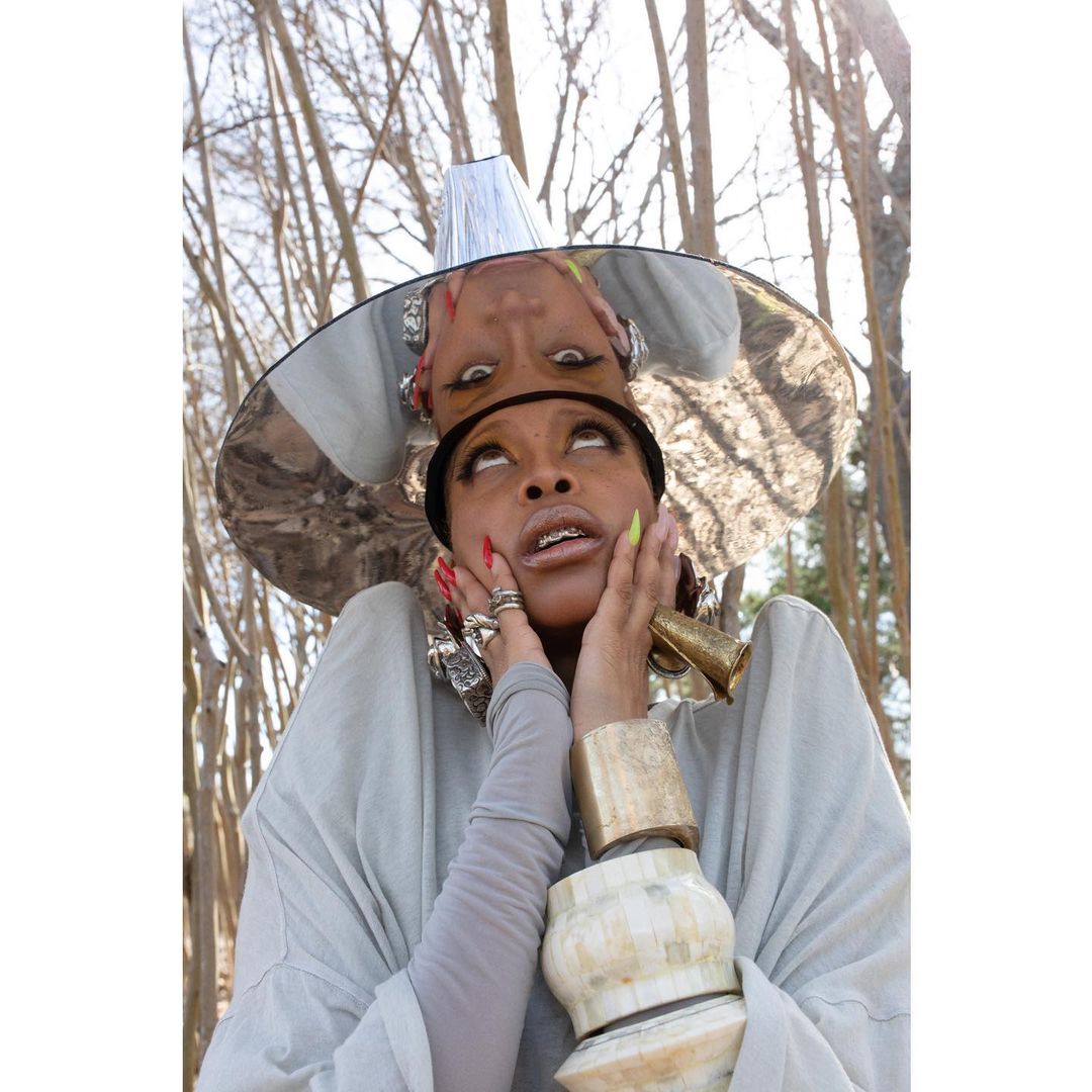 Portrait of an African-American woman with a silver hat looking up, wearing jewelry and light grey clothing in an outdoor setting.