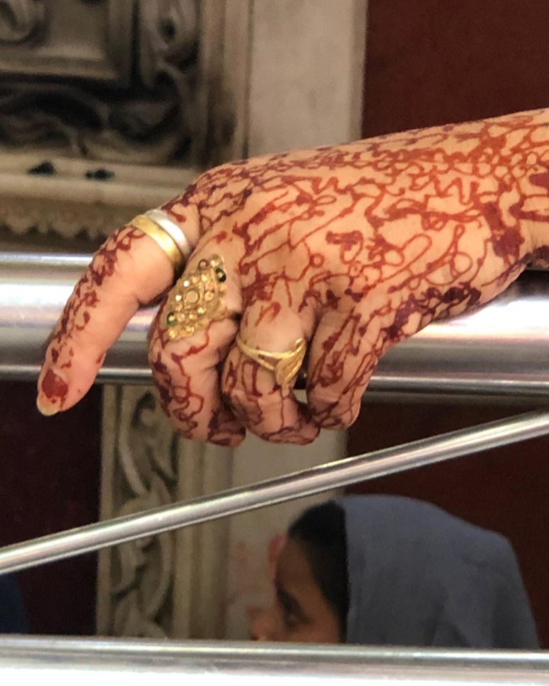 Close-up of an adult female hand with henna tattoos and gold rings, the background is blurred and suggests an indoor setting.
