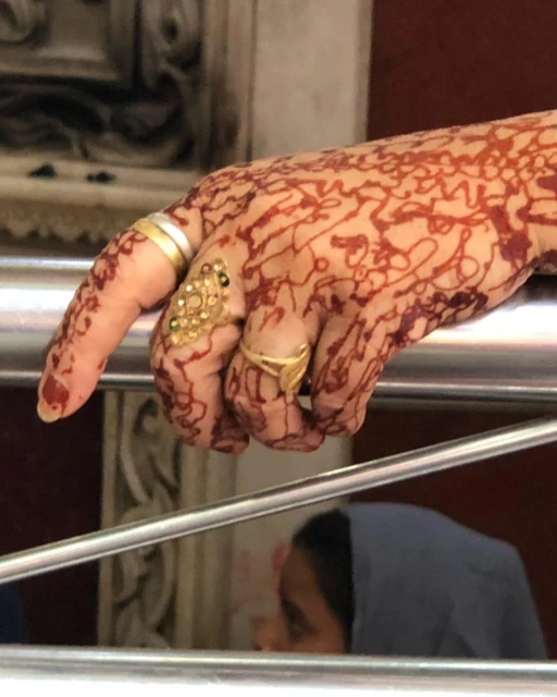 Close-up of an adult female hand with henna tattoos and gold rings, the background is blurred and suggests an indoor setting.