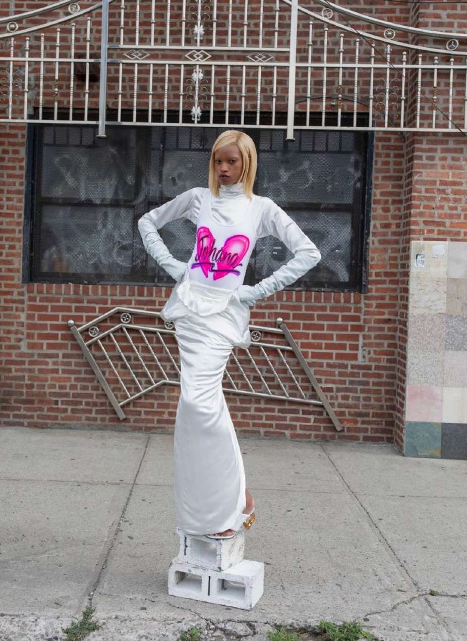 A tall, fair-skinned woman with blonde hair poses on concrete blocks outside, wearing a long white dress with a pink heart design.