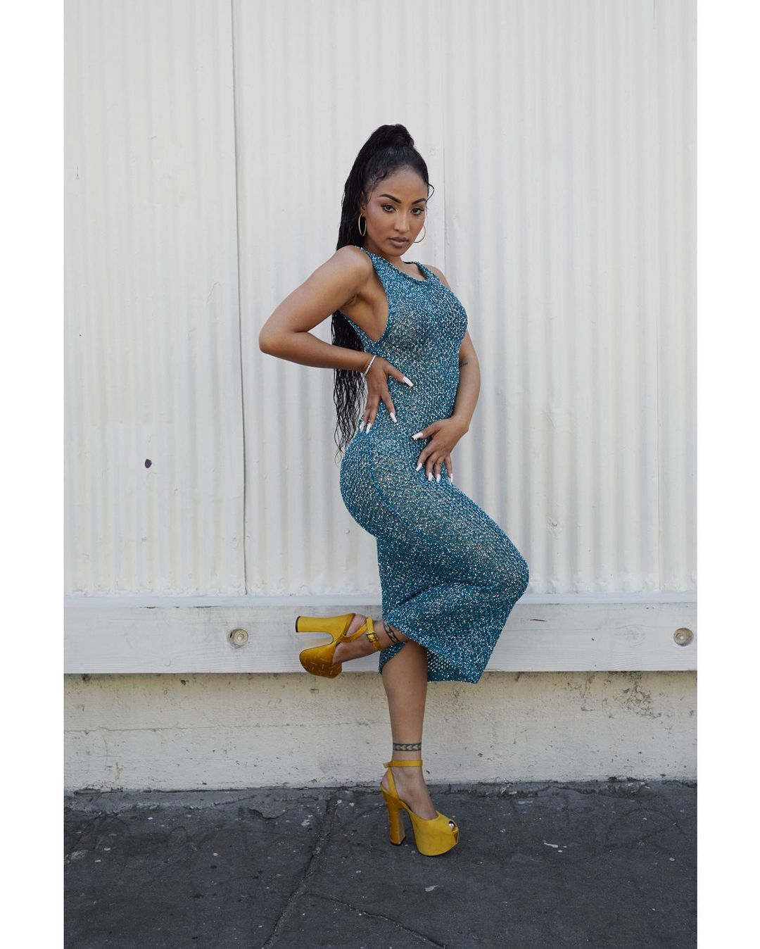 Attractive woman posing in an outdoor setting wearing a teal dress and bright yellow platform heels, with a neutral white backdrop.