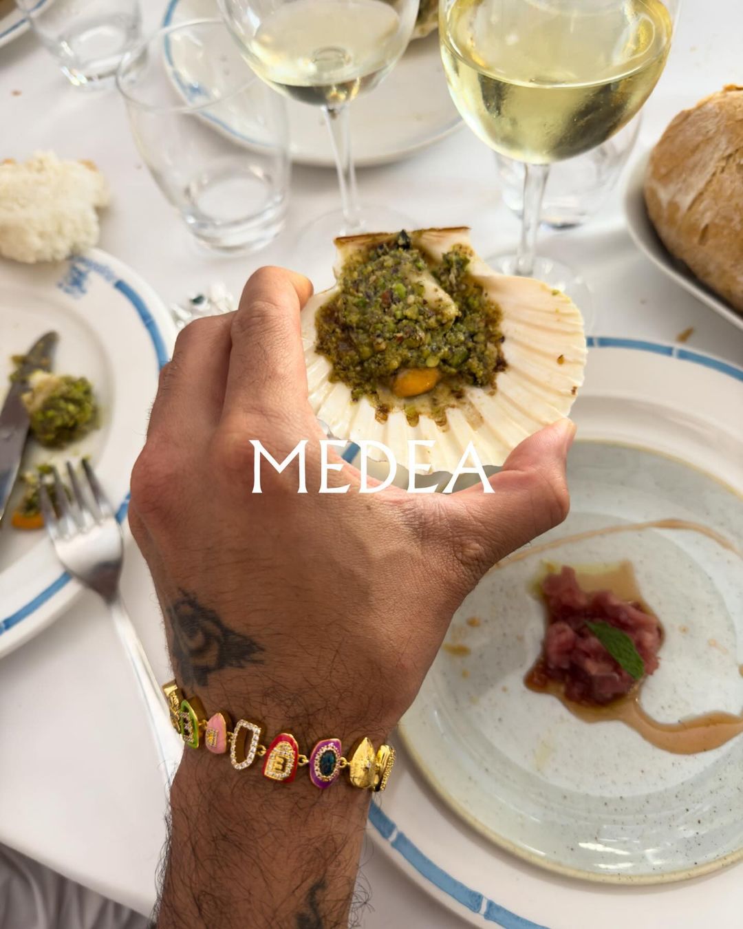 A person holds a scallop shell filled with food while seated at a table with wine and other dishes, showcasing a bracelet.