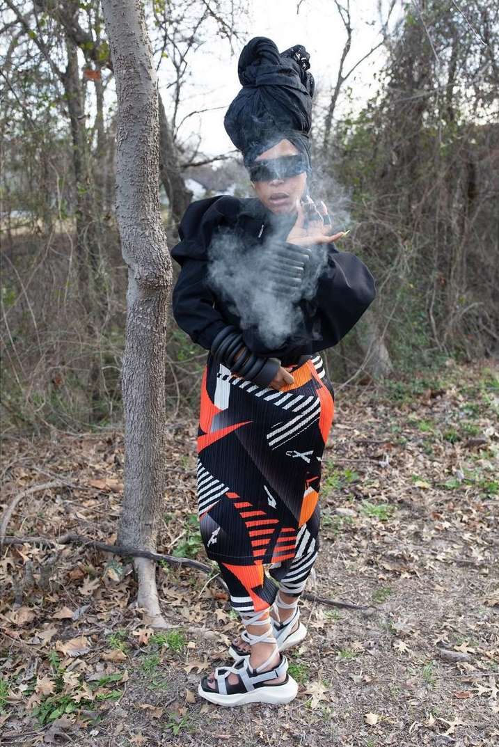Woman in streetwear fashion with smoke effect in outdoor wooded environment. Person in black top, patterned leggings, and sandals.