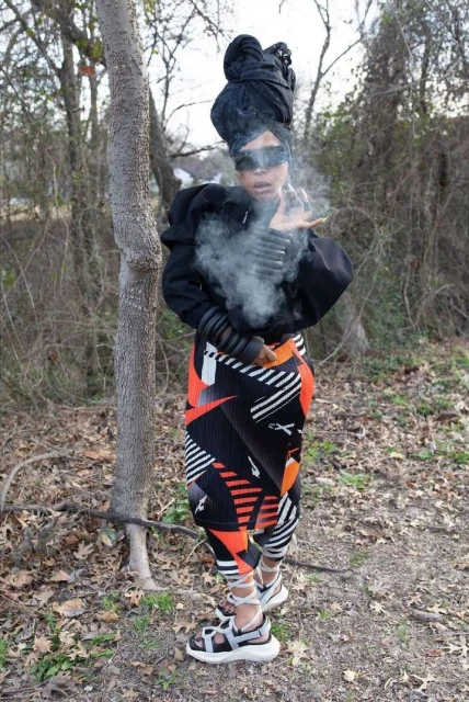 Woman in streetwear fashion with smoke effect in outdoor wooded environment. Person in black top, patterned leggings, and sandals.