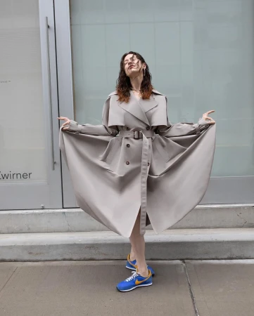 Woman in trench coat standing against building in blue and gold sneakers. Outdoor shot. Fashion editorial. Hair styled. 