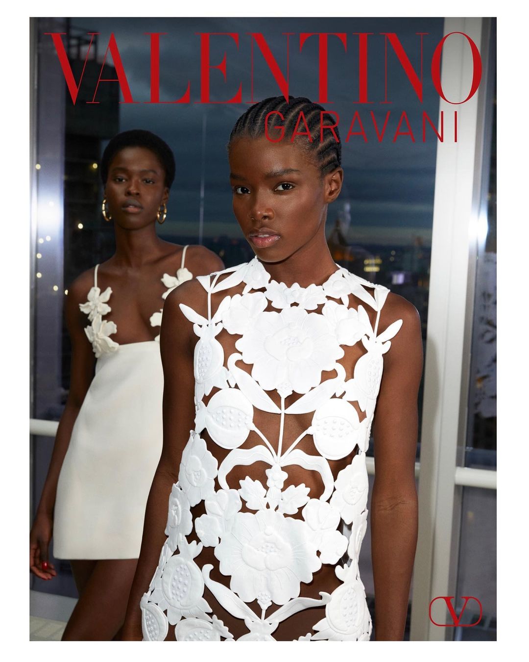 Two Black female fashion models showcasing white floral applique dresses indoors near a window with city lights in the background.