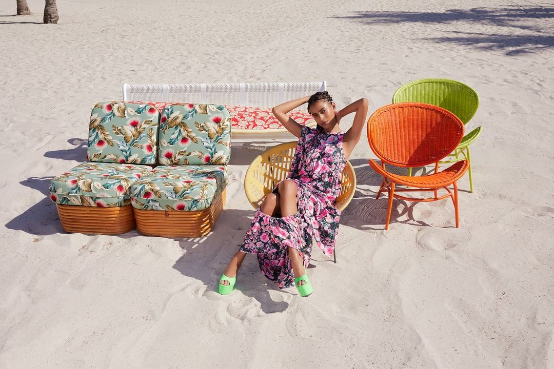 A woman in a floral dress sits on a yellow wicker chair on a beach, surrounded by colorful wicker furniture, under bright sunlight.