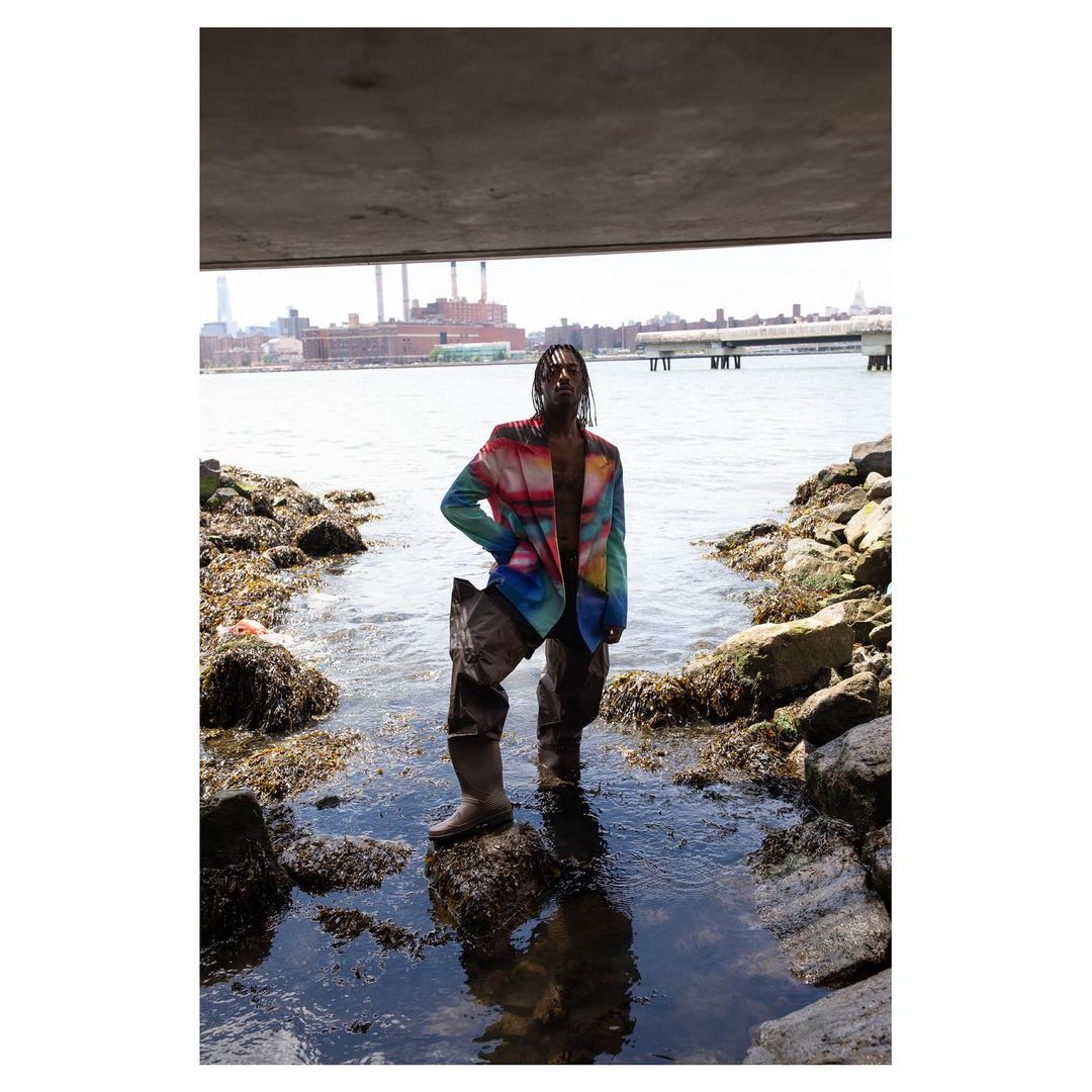 Man in colorful blazer and waders stands on rocks in a river under a bridge, with an industrial cityscape in the background.