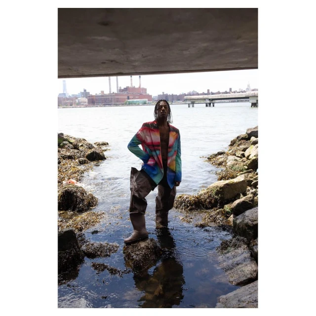 Man in colorful blazer and waders stands on rocks in a river under a bridge, with an industrial cityscape in the background.
