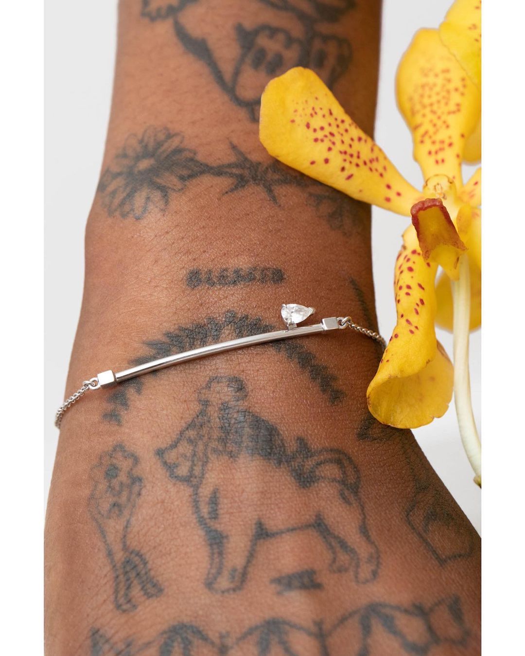 Close-up of a silver bracelet with a small diamond on a tattooed arm, with a yellow orchid flower in the background. Beauty shot.