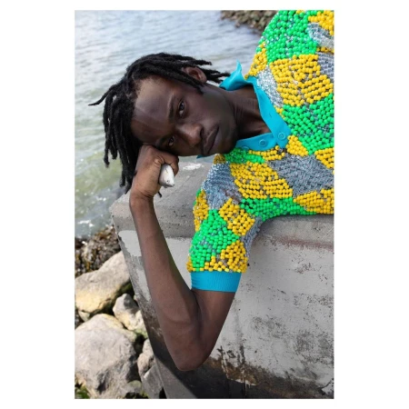 Man with dreadlocks wearing a beaded polo shirt leans against concrete near water with rocks in an outdoor setting during the daytime.