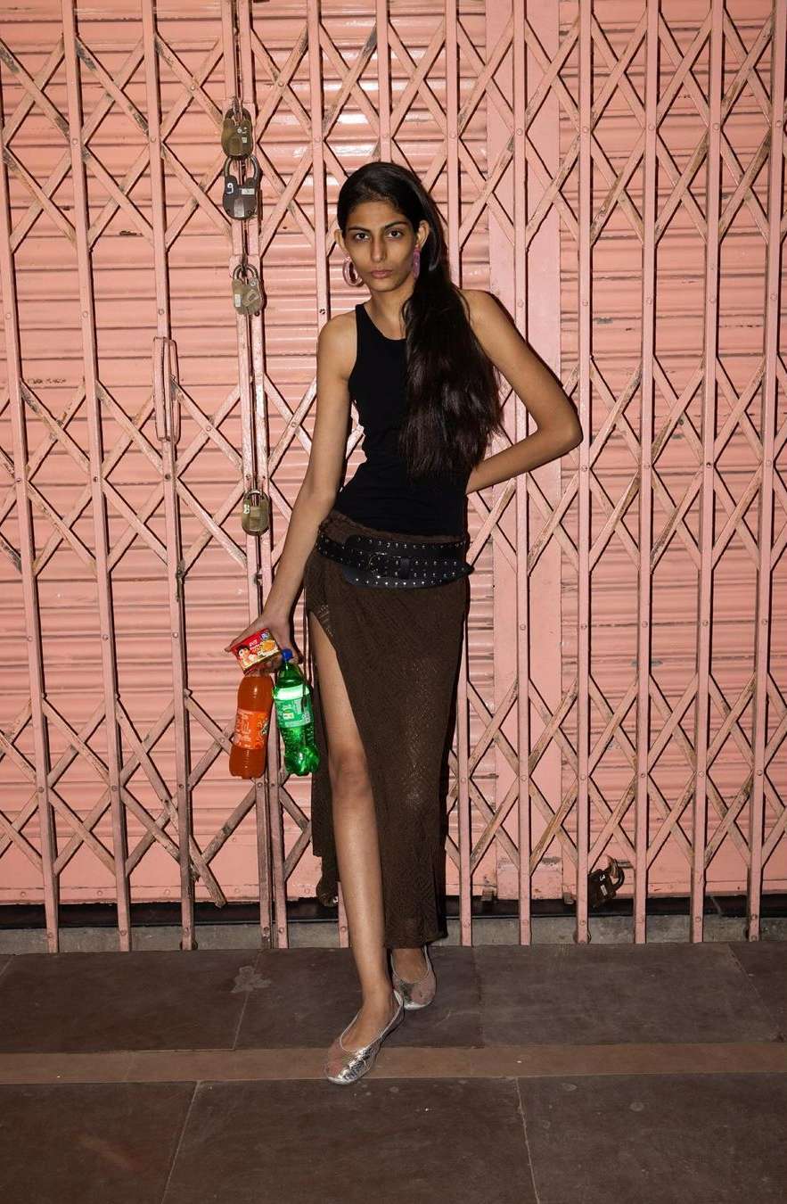 Young adult woman in a black top and brown skirt holding bottles poses in front of a pink metal gate in an urban environment.