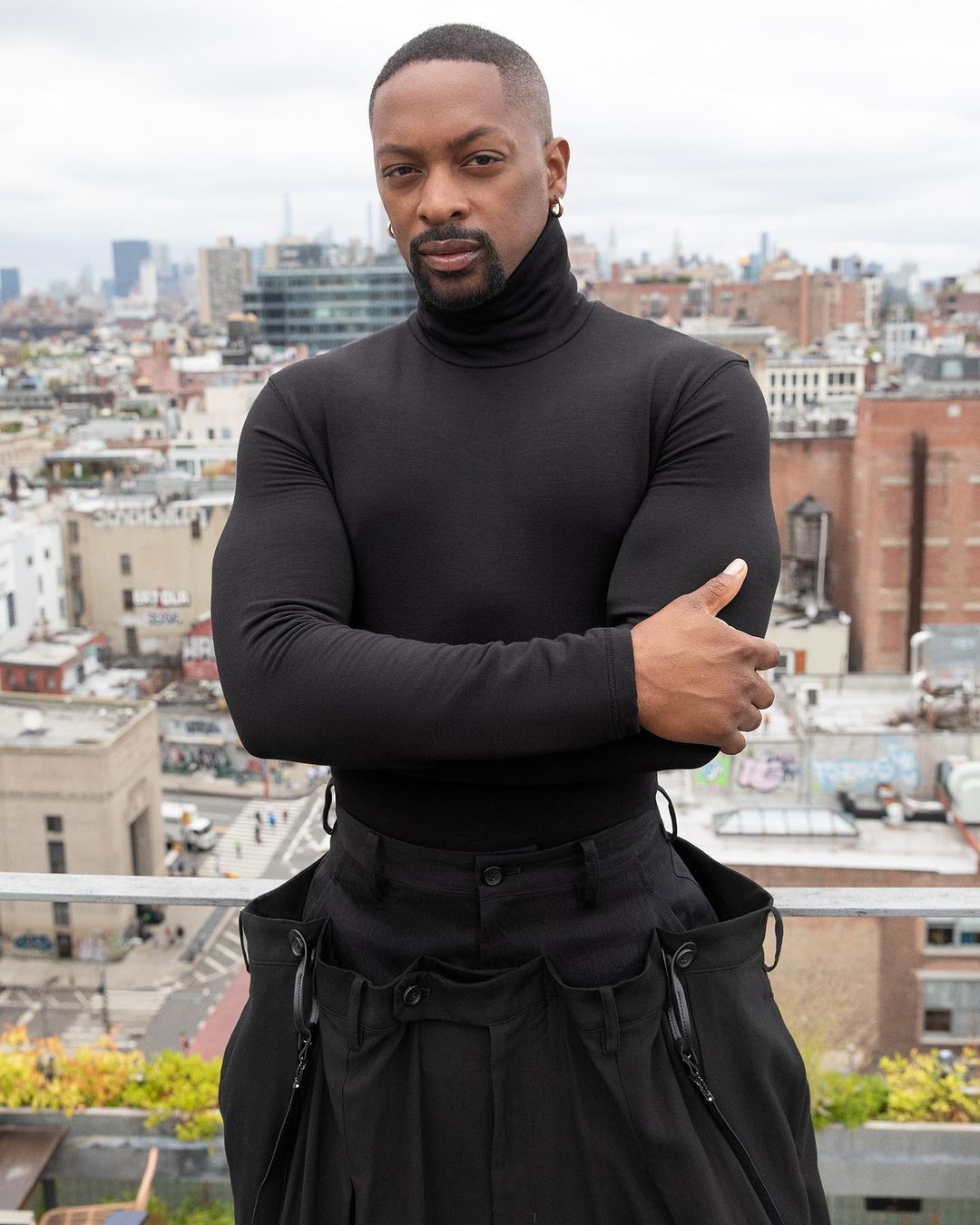 Portrait of an adult Black man outdoors with city skyline. He is wearing a black turtleneck and black pants with a serious facial expression.